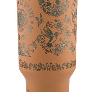 May include: A peach-colored tumbler with a silver floral and butterfly design. The tumbler has the words "BE BRAVE BE YOU" in a circular pattern. It has a white lid and a clear straw.