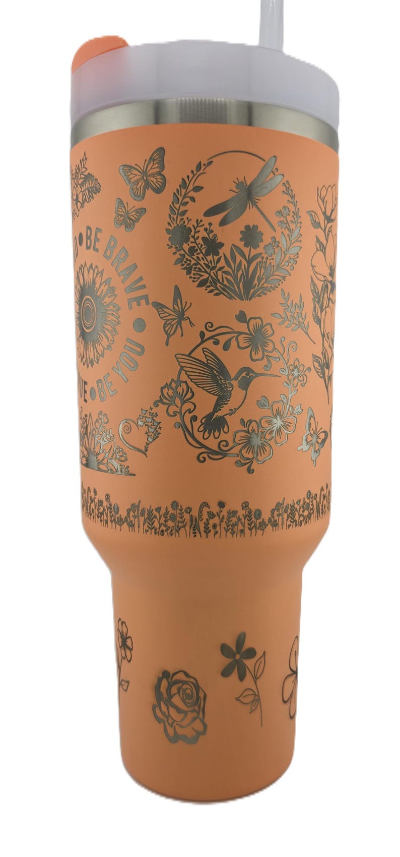 May include: A peach-colored tumbler with a silver floral and butterfly design. The tumbler has the words "BE BRAVE BE YOU" in a circular pattern. It has a white lid and a clear straw.