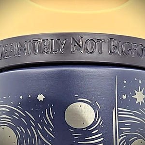 Pu&ograve; includere: Una tazza da viaggio blu scuro con un design a cielo notturno con stelle e lune. La tazza ha la scritta "DEFINITELY NOT BIGFOOT" in rilievo sulla parte superiore. Il coperchio &egrave; di colore giallo chiaro.