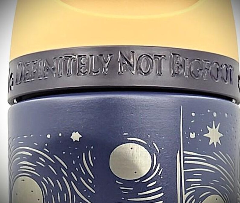 Pu&ograve; includere: Una tazza da viaggio blu scuro con un design a cielo notturno con stelle e lune. La tazza ha la scritta "DEFINITELY NOT BIGFOOT" in rilievo sulla parte superiore. Il coperchio &egrave; di colore giallo chiaro.