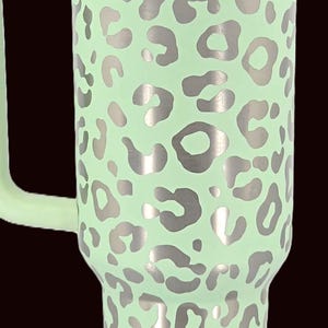 Puede incluir: Un vaso t&eacute;rmico grande, verde menta, con asa, con un estampado de leopardo plateado. El vaso tiene una tapa plateada y un asa verde menta. El logotipo de Stanley es visible en la parte inferior.