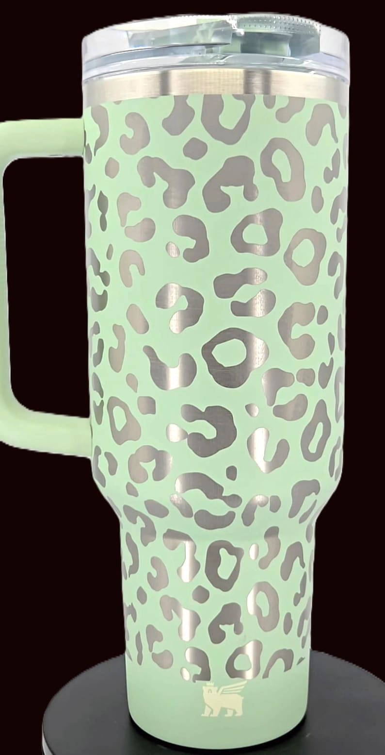 Puede incluir: Un vaso t&eacute;rmico grande, verde menta, con asa, con un estampado de leopardo plateado. El vaso tiene una tapa plateada y un asa verde menta. El logotipo de Stanley es visible en la parte inferior.