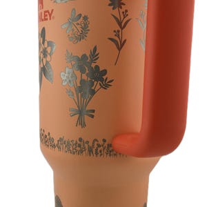 May include: A peach-colored insulated tumbler with a white lid and orange handle. The tumbler is decorated with silver floral designs and the brand name "Stanley". The handle is a vibrant orange color.