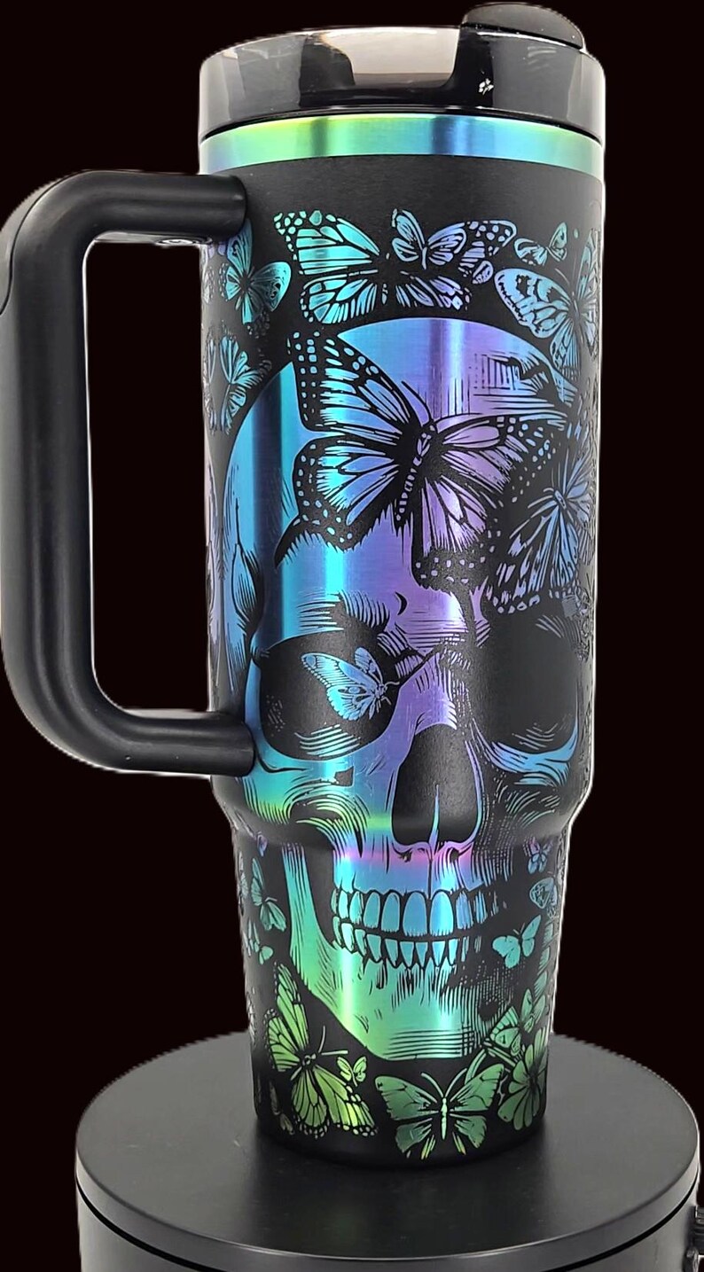 May include: Black travel mug with a skull and butterfly design. The skull is overlaid with iridescent blue, purple, and green butterflies. The mug has a black handle and a black lid with a metallic iridescent band.