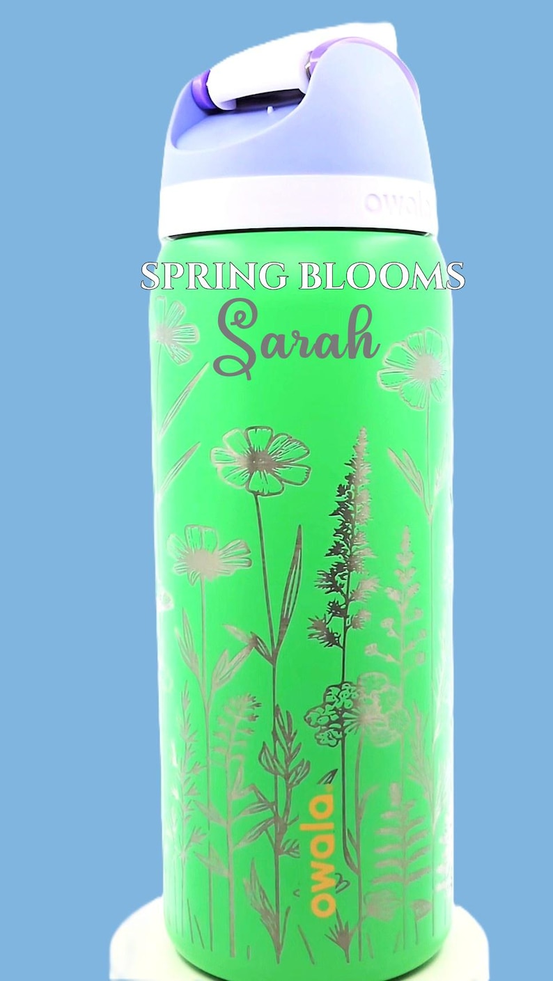 Pu&ograve; includere: Una borraccia Owala verde con coperchio e manico bianchi. La bottiglia &egrave; decorata con illustrazioni floreali argentate e le parole "Spring Blooms" e "Sarah". La bottiglia ha una capacit&agrave; di circa 710 ml.