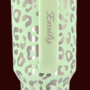 Puede incluir: Un vaso de color verde menta con un dise&ntilde;o de estampado de leopardo plateado. El vaso tiene un asa verde menta a juego con el nombre "Emily" impreso verticalmente. El vaso est&aacute; dise&ntilde;ado para contener bebidas.