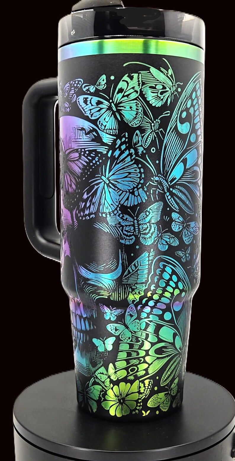 May include: Black stainless steel tumbler with a wraparound design of butterflies in shades of blue, green, and purple. The tumbler has a black handle and a black lid with a colorful iridescent trim. The design includes a skull.