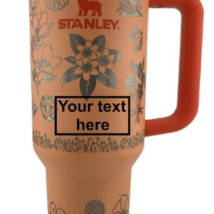 May include: A peach-colored Stanley tumbler with a silver floral design and the text "Your text here". It has a silver top, an orange handle, and the Stanley logo. The tumbler is designed to hold approximately 30 ounces.
