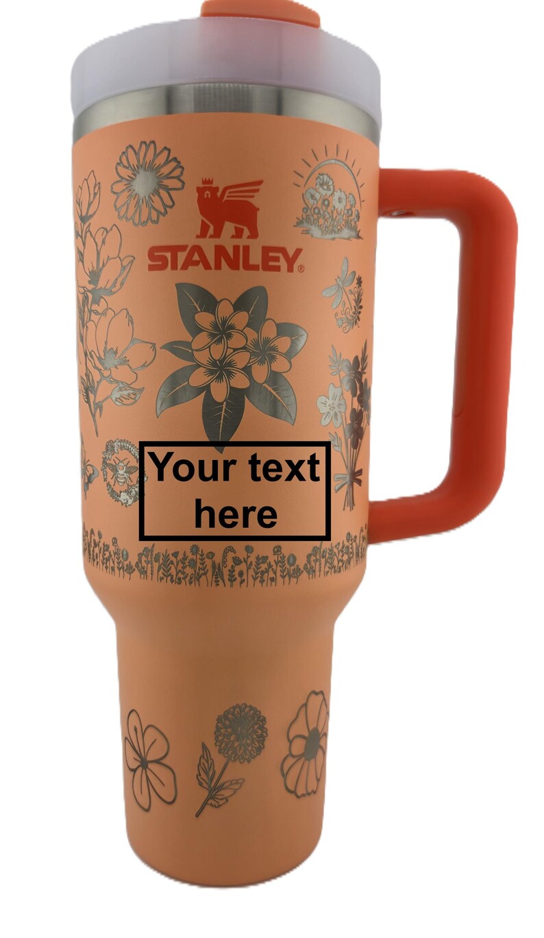May include: A peach-colored Stanley tumbler with a silver floral design and the text "Your text here". It has a silver top, an orange handle, and the Stanley logo. The tumbler is designed to hold approximately 30 ounces.