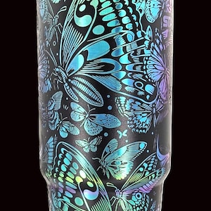 May include: Black tumbler with a vibrant butterfly design. The butterflies are in shades of blue, green, and purple, creating a colorful contrast against the black background. The tumbler has a sleek, modern design.