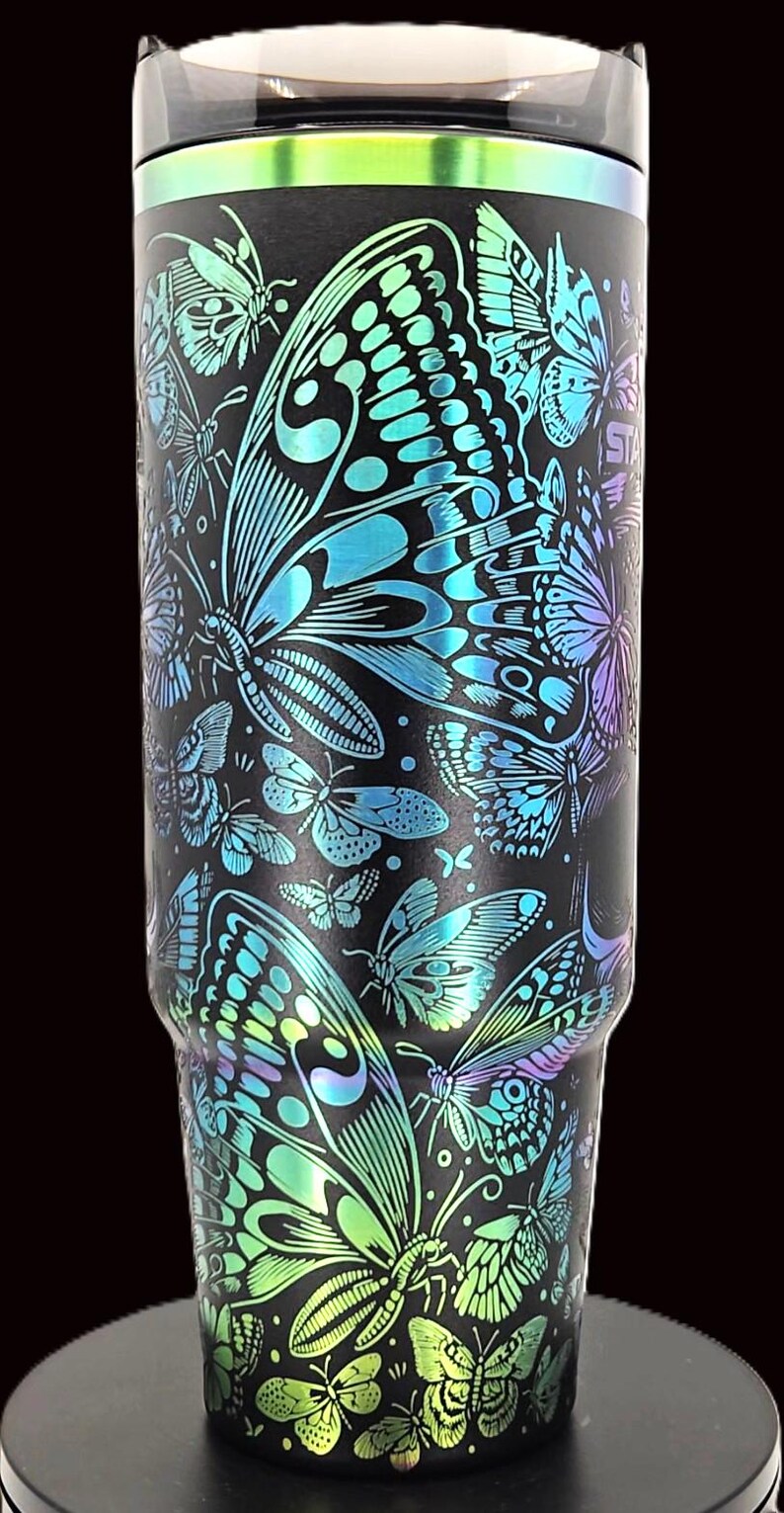 May include: Black tumbler with a vibrant butterfly design. The butterflies are in shades of blue, green, and purple, creating a colorful contrast against the black background. The tumbler has a sleek, modern design.
