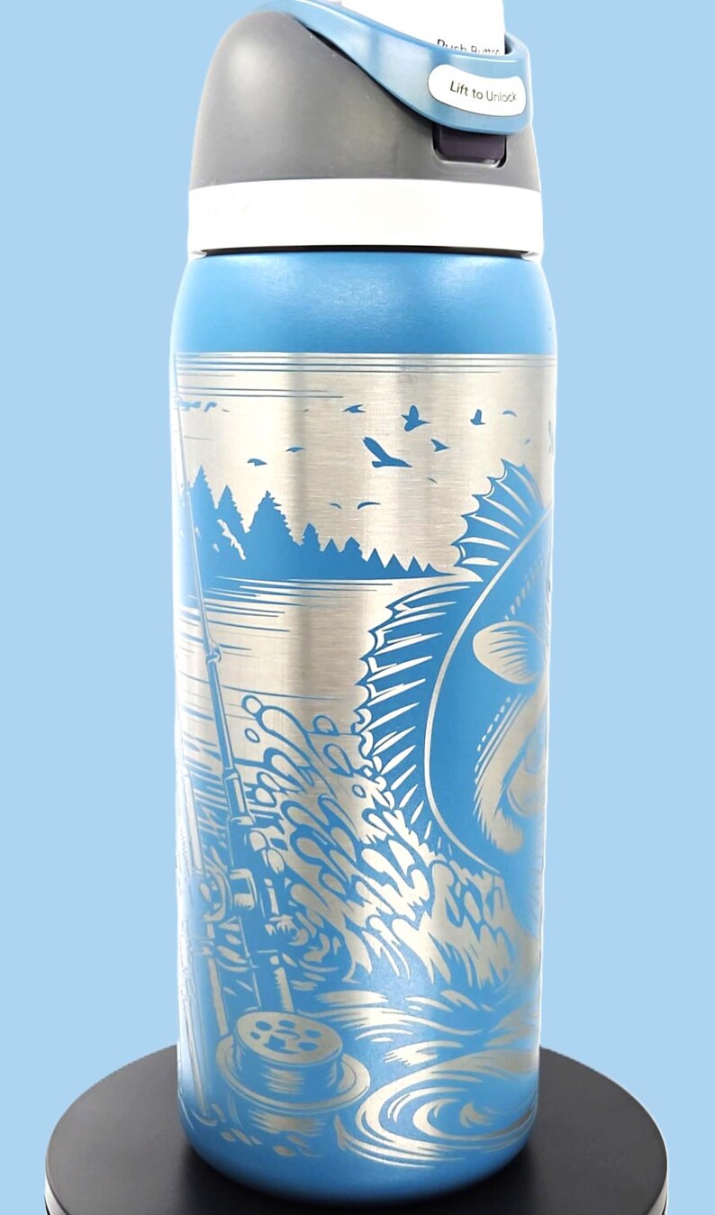 May include: Blue water bottle with a silver fishing scene design. The illustration features a fish jumping out of the water, a fishing rod, and a lake with trees and birds. The lid is gray and white.