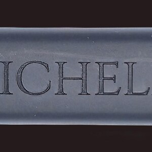 May include: A rectangular, dark gray object with rounded edges. The word "MICHELE" is engraved in a serif font across the center. The left side has a dotted texture.