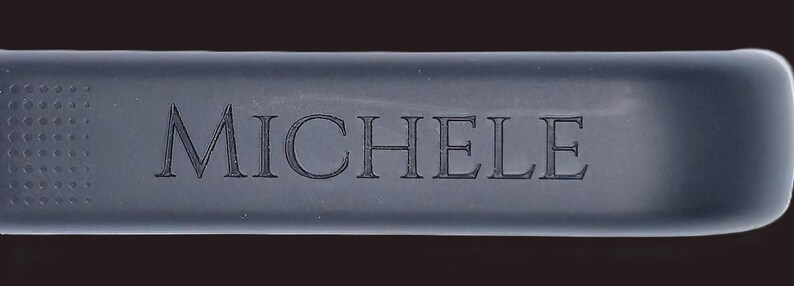 May include: A rectangular, dark gray object with rounded edges. The word "MICHELE" is engraved in a serif font across the center. The left side has a dotted texture.