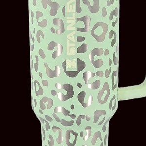 Puede incluir: Un vaso Stanley verde menta con un estampado de leopardo plateado. El vaso tiene un asa y una tapa transparente. El logotipo de Stanley es visible en el lateral. Este termo aislado est&aacute; dise&ntilde;ado para mantener las bebidas fr&iacute;as o calientes.