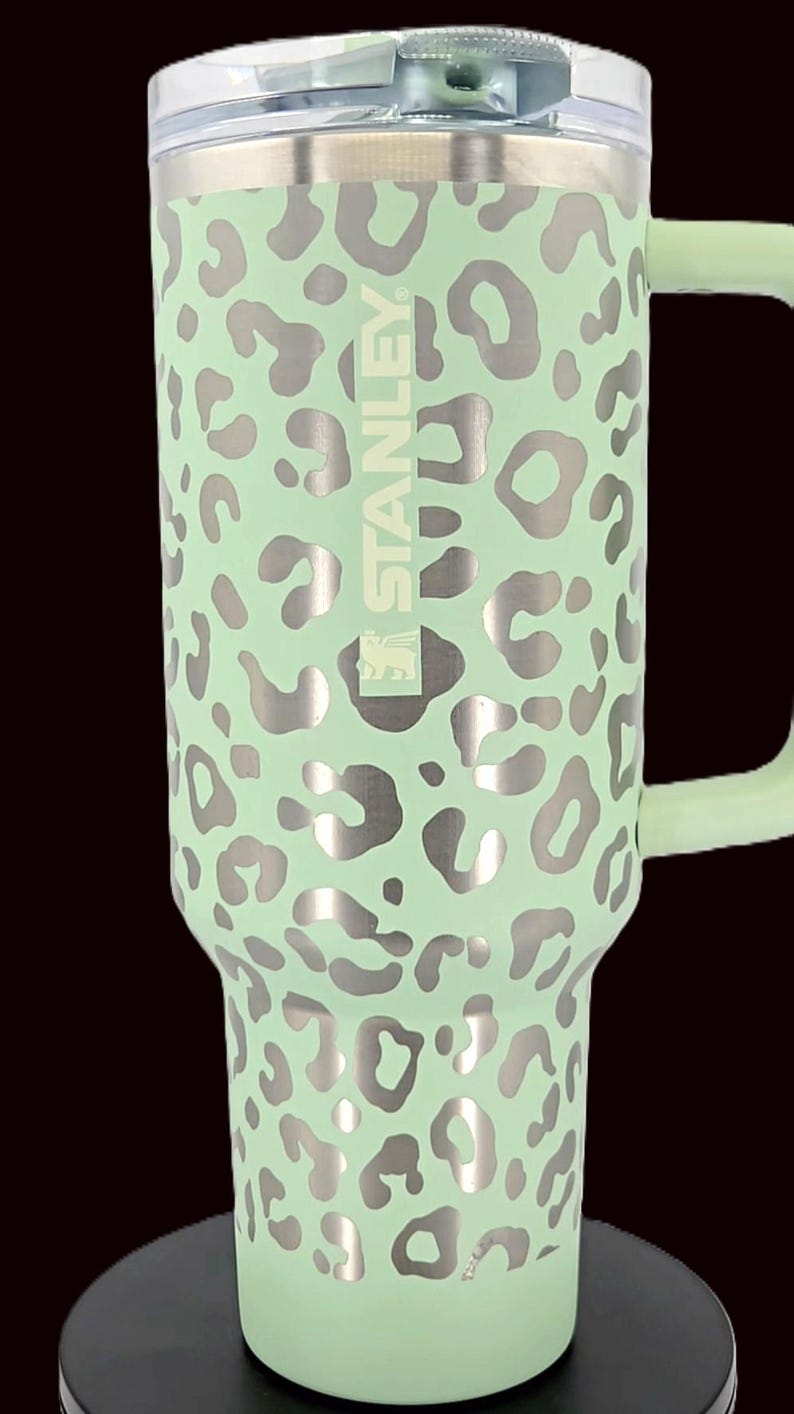 Puede incluir: Un vaso Stanley verde menta con un estampado de leopardo plateado. El vaso tiene un asa y una tapa transparente. El logotipo de Stanley es visible en el lateral. Este termo aislado est&aacute; dise&ntilde;ado para mantener las bebidas fr&iacute;as o calientes.