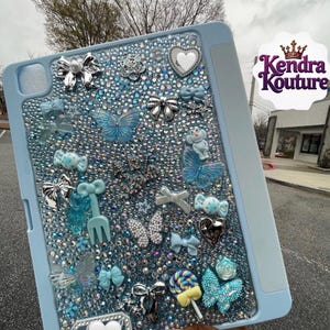 May include: A light blue tablet case covered in small iridescent crystals and various charms including silver bows, blue butterflies, candy shapes, hearts, and a lollipop. The text Kendra Kouture is visible in the upper right corner of the image.