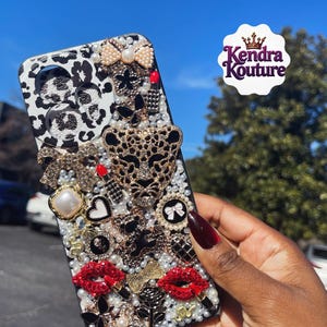 May include: A bedazzled phone case with a black and white leopard print design. The case is adorned with pearls, bows, a gold leopard head, red lips, and other decorative elements. The Kendra Kouture logo is visible.
