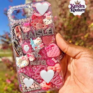 May include: A clear phone case decorated with pink and silver glitter, rhinestones, and various charms. Charms include Hello Kitty figures, hearts, bows, and the name "Kaislei". The case has a decorative border of rhinestones.