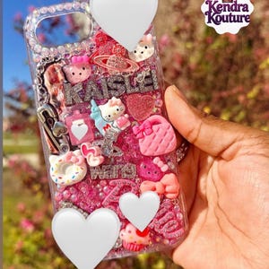 May include: A clear phone case decorated with pink and white charms, including miniature figures of a cat, a handbag, a cupcake, and a doughnut. The case features silver glitter, rhinestones, and the name KAISLEI spelled out in silver letters.