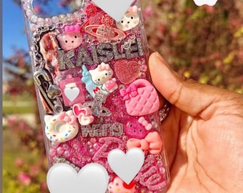 May include: A clear phone case decorated with pink and white charms, including miniature figures of a cat, a handbag, a cupcake, and a donut. The case features silver glitter, rhinestones, and the name KAISLEI spelled out in silver letters.