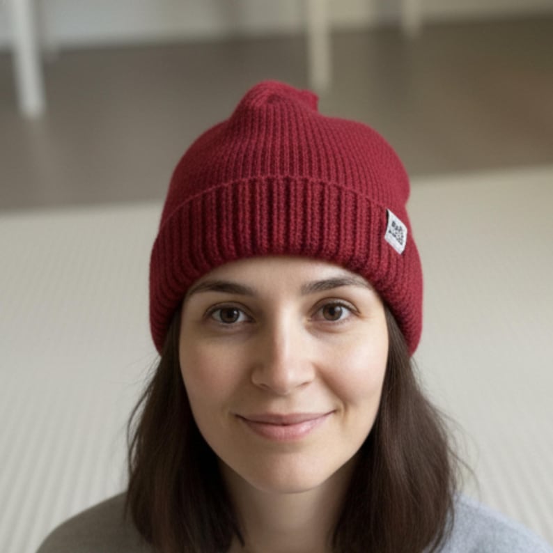 K&ouml;nnte beinhalten: Burgunderfarbene Strickm&uuml;tze mit Rippenstruktur. Der Hut hat ein kleines wei&szlig;es Etikett an der Seite. Die Person, die die M&uuml;tze tr&auml;gt, hat braune Haare und ein graues Oberteil.