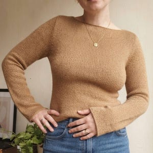 May include: A long-sleeved, tan-colored knit sweater featuring a wide boat neckline and a fine, textured stitch pattern. The sweater is paired with blue denim jeans, creating a simple and classic look that highlights the warm, neutral tone.