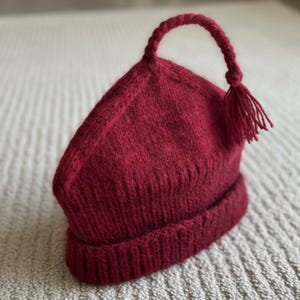 K&ouml;nnte beinhalten: Eine weinrote Strickm&uuml;tze mit einem einzigartigen Design. Die M&uuml;tze hat einen gerippten Saum und eine spitz zulaufende Oberseite mit einem geflochtenen Griff und Quaste. Die M&uuml;tze ist aus weichem, strukturiertem Garn gefertigt.