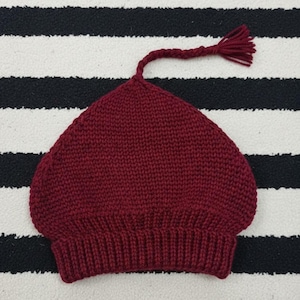 Könnte beinhalten: Eine bordeauxfarbene Strickmütze mit abgerundeter Krone und geripptem Bund. Ein geflochtener Kordelzug erstreckt sich von der Oberseite der Mütze und endet in einer Quaste. Die Mütze ist vor einem schwarz-weiß gestreiften Hintergrund abgebildet.