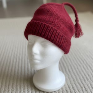 May include: A knitted burgundy beanie hat with a long, curved tassel. The hat is displayed on a white mannequin head. The hat's brim is ribbed, and the tassel hangs down from the top. The hat is made of a soft, textured material.