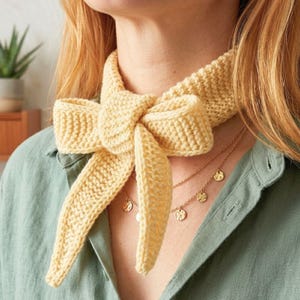 Peut inclure: Un foulard tricoté jaune pâle présentant un motif de mailles texturé et un grand nœud noué sur le devant. Le foulard est porté autour du cou, reposant sur un collier superposé doré délicat avec de petites breloques métalliques martelées.