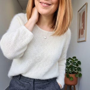 Può includere: Un maglione in maglia a maniche lunghe color crema con scollo rotondo e consistenza soffice, abbinato a pantaloni grigio scuro. Una delicata collana a catena argentata con un singolo ciondolo a perla bianca poggia sullo scollo del maglione.