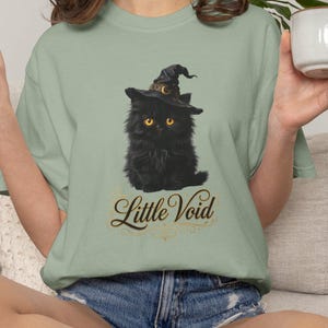 Può includere: Una t-shirt verde salvia con la grafica di un gatto nero soffice che indossa un cappello da strega a punta e ha occhi gialli. Sotto il gatto, il testo Little Void è stampato in un carattere dorato decorativo. Maglietta a maniche corte.