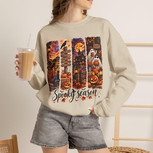 Op de afbeelding: Een beige sweater met ronde hals en een kleurrijke grafische print van Halloween-taferelen, waaronder pompoenen, geesten, een spookhuis en een wegwijzer met de tekst Spooky season in een cursief lettertype onderaan het ontwerp.