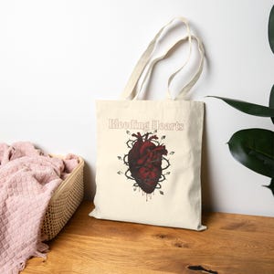 May include: Cream-colored tote bag featuring a dark red anatomical heart design with thorns and the text "Bleeding Hearts". The heart has dripping blood details. The bag has long handles.