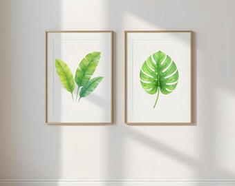 May include: Two framed botanical prints with watercolour illustrations of green leaves. One print shows three banana leaves, the other a monstera leaf. The frames have a gold finish. The background is white.