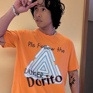 May include: Orange t-shirt with the text "Pls Follow the DANGER Dorito" and a graphic of a triangle. The person is wearing colorful patterned pants and a silver bracelet and watch.