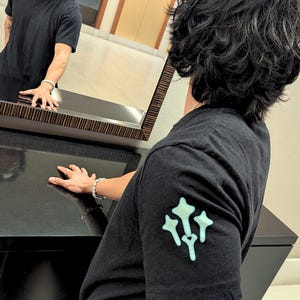 Puede incluir: Camiseta negra de manga corta con un gráfico verde claro en la manga izquierda. La persona de la imagen lleva una pulsera y gafas. La persona se refleja en un espejo.