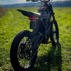 Könnte beinhalten: Schwarzes Elektro-Dirtbike mit Stollenreifen, einem kleinen Sitz und einem Rücklicht. Das Motorrad steht auf einer Wiese unter blauem Himmel mit Wolken. Der Lenker ist sichtbar.