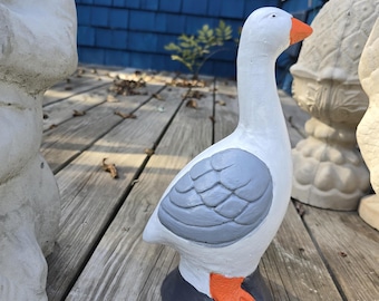 May include: A white goose statue with gray wings and orange feet and beak. The decorative garden ornament is standing on a dark gray base, set against a weathered wooden deck and a blue house.