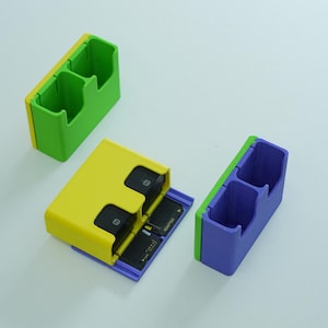 May include: Three colorful SD card holders in green, yellow, and blue. The yellow holder is open, revealing several black SD cards. The other two holders are closed, showing their dual-compartment design. The holders are made of plastic.