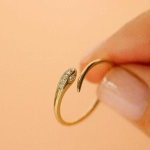 May include: A gold-colored ring with a snake-like design. The ring features a curved shape with a head-like end adorned with small, clear gemstones. The ring is held against a peach-colored background.