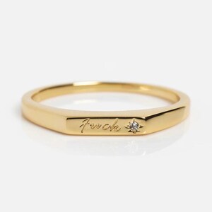 May include: A gold-colored ring with the word "fuck" engraved on the front. A small, clear gemstone is set next to the word. The ring has a geometric, rectangular design.