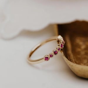 May include: A gold band ring featuring a series of small, round, ruby-colored gemstones. The gemstones are set in individual bezels along the top of the ring. The ring is displayed on a white surface.