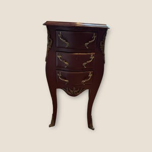May include: A small, antique-style chest of drawers with three curved drawers. The chest is a deep burgundy color with ornate gold-colored hardware and accents. The legs are slender and curved, adding to the vintage aesthetic.