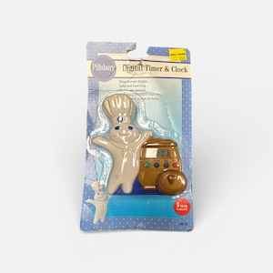 May include: A Pillsbury digital timer and clock in its original packaging. The timer is brown and shaped like a vintage oven, with the Pillsbury Doughboy figure. The packaging is blue with white polka dots and includes the text "Digital Timer & Clock".