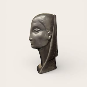 Puede incluir: Una escultura de bronce oscuro de la cabeza de una mujer de perfil. La escultura presenta un diseño suave y estilizado con un tocado drapeado. La cabeza está colocada sobre un cuello corto, y la estética general recuerda al arte clásico.