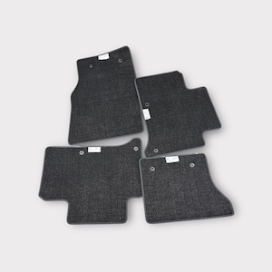 Puede incluir: Cuatro alfombrillas de coche de color gris oscuro. Las alfombrillas están hechas de un material texturizado y tienen forma rectangular con bordes redondeados. Cada alfombrilla tiene una pequeña etiqueta blanca y un agujero para sujetarlas.