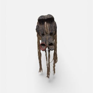 May include: A weathered, dark-colored mask with a stylized face, featuring two red-rimmed eye holes and long, dangling strands of natural fiber. The mask has a rough texture and appears to be handmade, possibly for ceremonial or decorative purposes.
