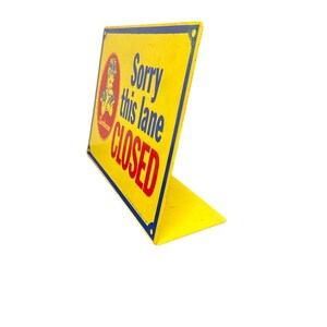 Op de afbeelding: Geel metalen bord met de tekst "Sorry this lane CLOSED" in blauw en rood. Het bord heeft een blauwe rand en een logo in de linkerbovenhoek. Het bord is schuin en heeft een standaard.